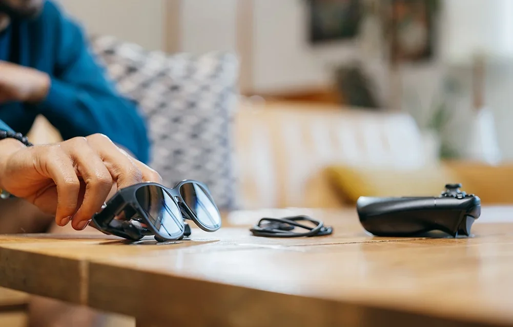 Ranka siekia juodų XR akinių, gulinčių ant medinio stalo šalia laido ir žaidimų valdiklio