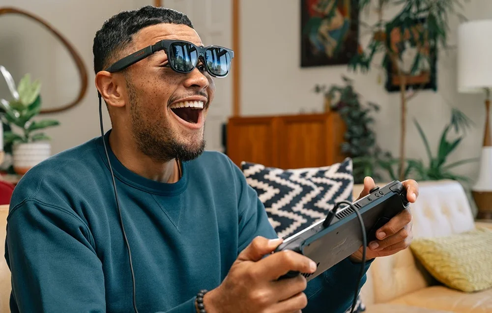 Vyras su XR akiniais juokiasi žaisdamas vaizdo žaidimus su valdikliu rankose svetainėje