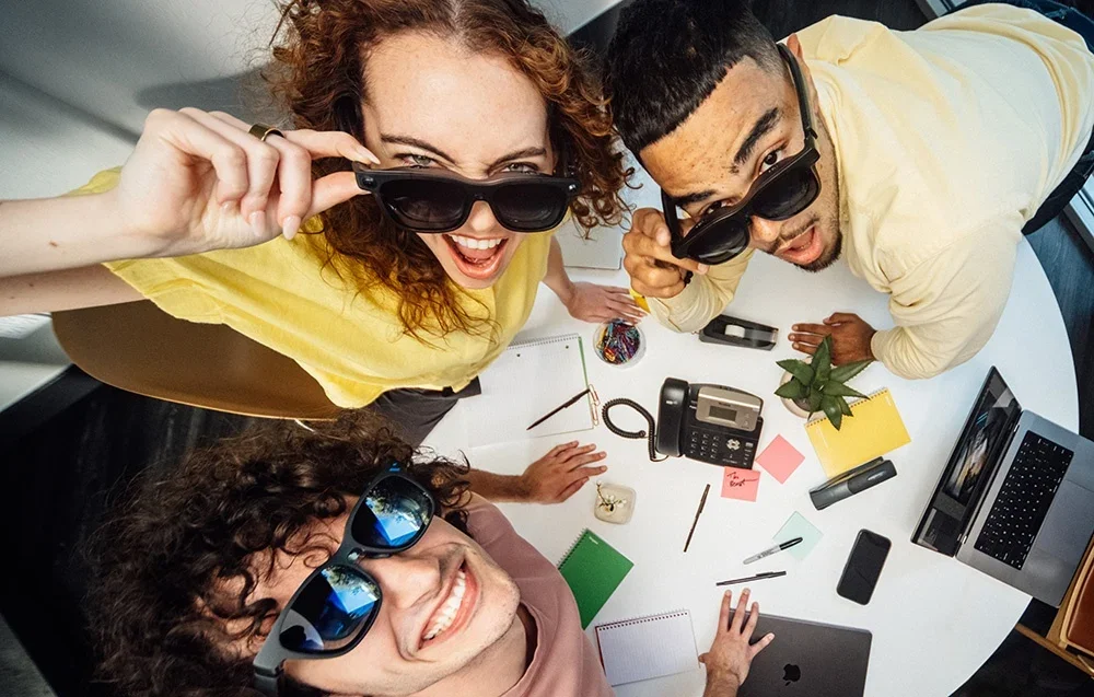 Trys darbuotojai su XR akiniais šypsosi prie stalo su nešiojamaisiais kompiuteriais ir lipniais lapeliais