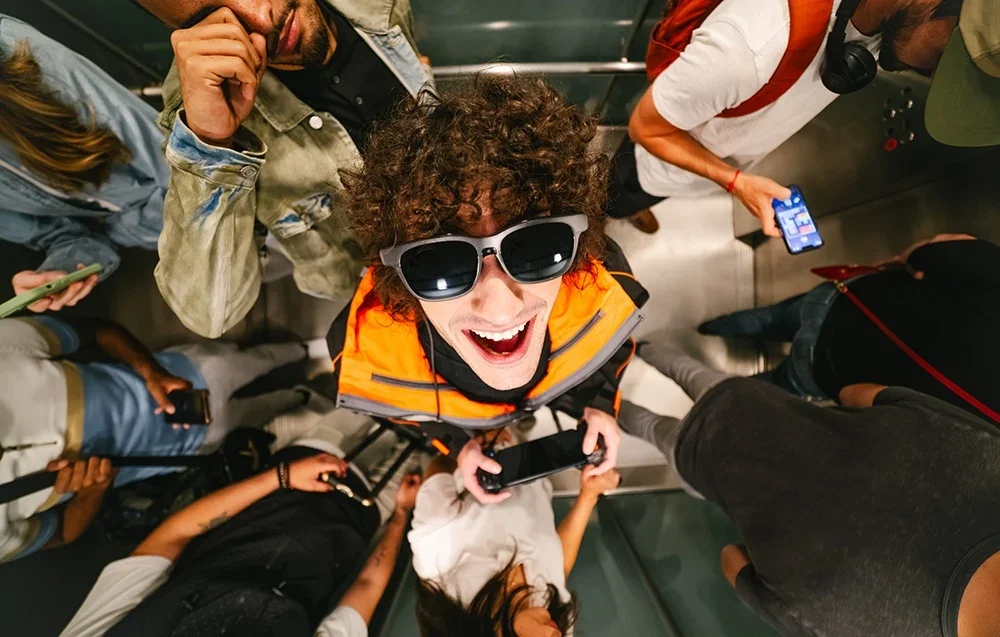 Žmogus oranžine liemene juokiasi dėvėdamas XR akinius, apsuptas kitų žmonių