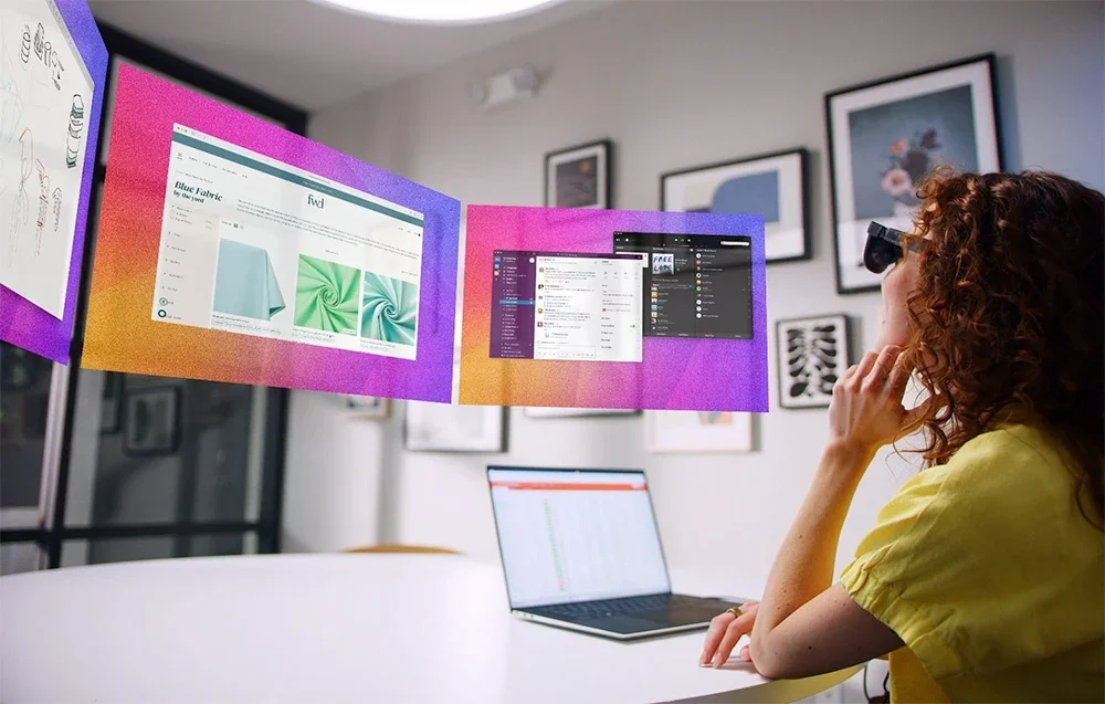 Moteris su XR akiniais dirba prie stalo su nešiojamuoju kompiuteriu, žiūrėdama virtualius ekranus ore