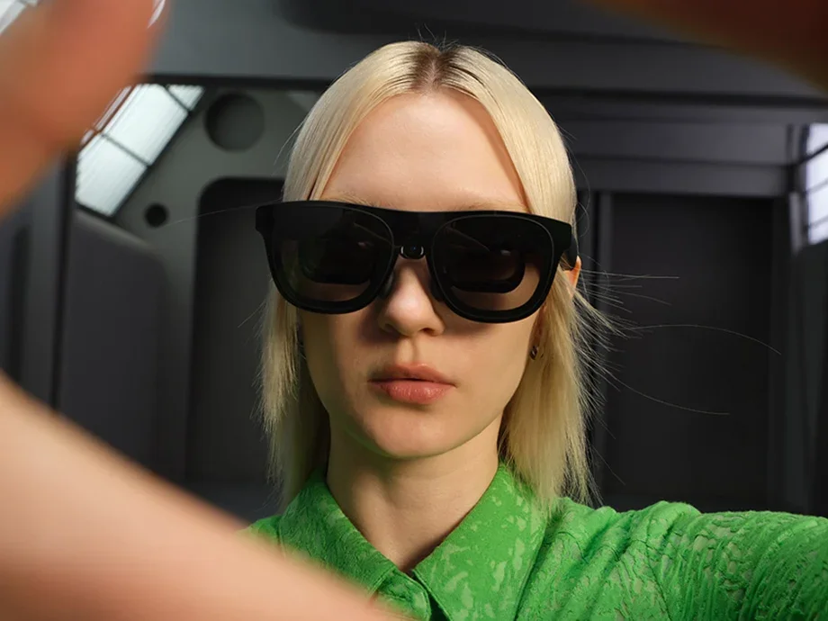 A woman in a green shirt looking ahead and wearing black AR glasses with a camera in the middle.